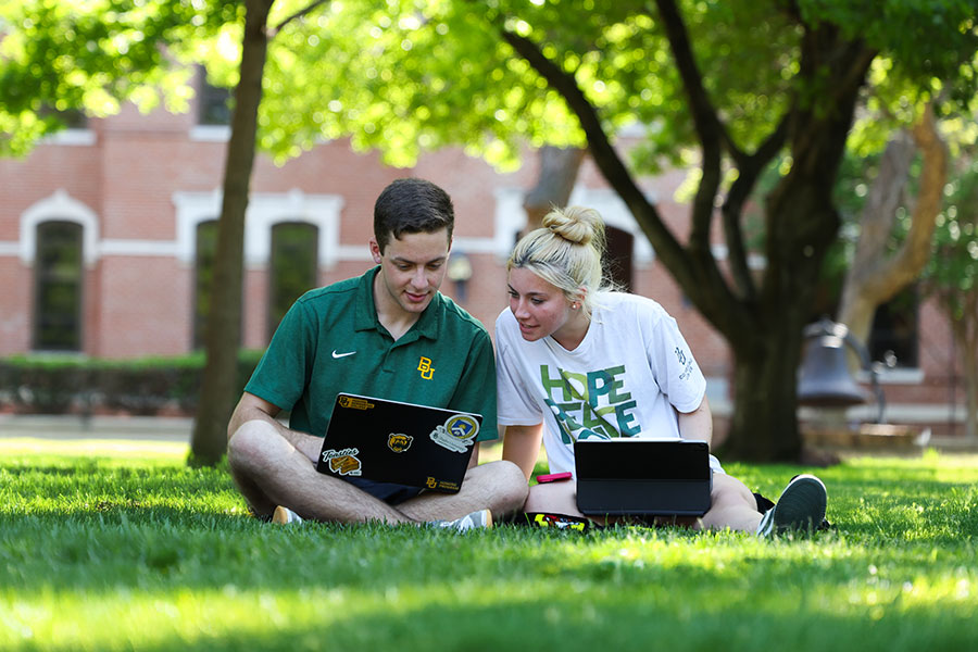 Two students on computers sitting on the grass on campus